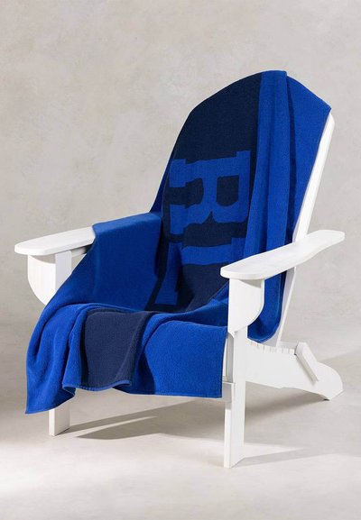 Λευκή ξύλινη πολυθρόνα με μια πετσέτα μπλε και ναυτικού χρώματος περασμένη πάνω της, με έντονα γράμματα σε ένα μινιμαλιστικό περιβάλλον.