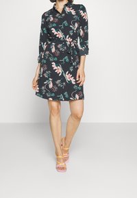Vestito floreale con sfondo scuro, caratterizzato da un colletto, maniche lunghe e una vita legata. Abbinato a sandali con cinturini multicolori.