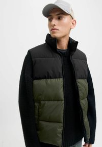 Crna i maslinasto zelena prsluk od puffer materijala s visokim ovratnikom, zatvaranjem na zip, i teksturiranom crnom pletenom džemperom ispod. Svijetlo siva kapa na glavi.