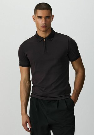 Mann mit kurzen Haaren, der ein schwarzes Kurzarm-Poloshirt mit einem Reißverschluss vorne und schwarze Hose trägt, steht mit einer Hand in der Tasche.