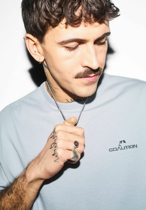 Mężczyzna z kręconymi włosami, wąsami i tatuażami na dłoniach, ubrany w szarą koszulkę Coalition, trzymający srebrny wisiorek na łańcuszku blisko klatki piersiowej.