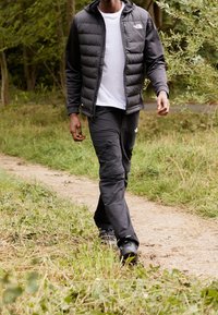 Sort pufferjakke med hætte, hvid t-shirt og sorte cargo-bukser, går på en sti omgivet af grønne omgivelser.