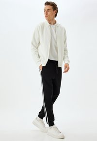 Chaqueta blanca con cremallera sobre una camiseta gris, combinada con pantalones de chándal negros con rayas laterales blancas y zapatillas blancas. Tela suave y casual.