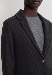 Pinstriped black blazer with a notched lapel, two buttons, and a textured gray sweater underneath. Fabric has a soft finish.