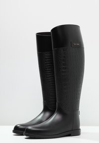 Botas negras hasta la rodilla con un acabado brillante, que presentan un diseño texturizado similar al cuero de cocodrilo en la parte superior y una suela de goma lisa.