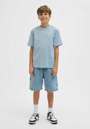 Boy standing and smiling, wearing a light blue t-shirt, denim shorts, white socks, and black-and-white sneakers, against a plain white background.