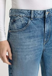Gros plan sur une personne portant un jean en denim bleu et une chemise à manches longues blanche, avec une main reposant sur son côté.