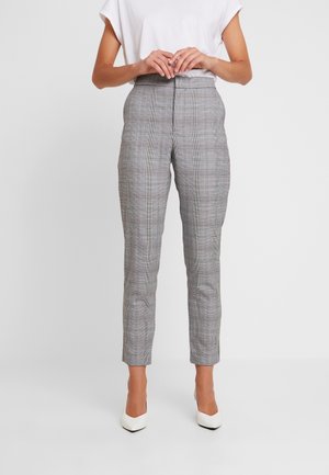 Person iført grå, ternede, højtaljede, slim-fit bukser, hvid ærmeløs top og hvide spidse hæle, stående med hænderne foldet.