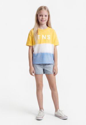 Junges Mädchen stehend, trägt ein gelb-weiß-blaues TNS-Farbblock-T-Shirt, helle Jeansshorts und weiße Turnschuhe vor weißem Hintergrund.