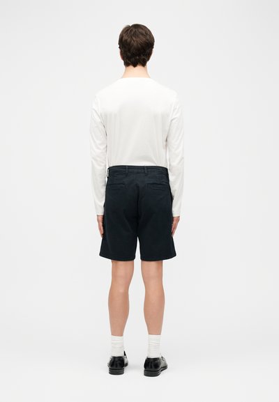 Άνδρας που στέκεται με την πλάτη γυρισμένη, φορώντας λευκό πουκάμισο με μακριά μανίκια, μαύρο σορτς μέχρι το γόνατο, λευκές κάλτσες και μαύρα επίσημα παπούτσια.