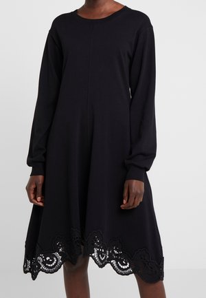 Langærmet sort kjole i blødt stof med en udvidet silhuet og detaljeret blondekant forneden. Enkel rund halsudskæring.