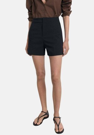 Pantaloni corti neri a vita alta con una texture liscia e un design sartoriale, abbinati a una camicia marrone con bottoni e sandali piatti neri.