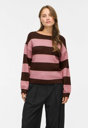 Femme portant un pull ample rayé rose et marron et un pantalon noir à jambes larges, debout sur fond blanc.