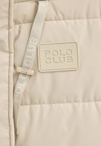 Beżowa puchowa kurtka o pikowanej fakturze, z prostokątną kremową naszywką z wytłoczonym napisem "POLO CLUB" oraz pasującym troczkiem.