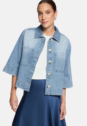 Žena v svetlomodrej rifľovej bunde s nášivkami a veľkými gombíkmi, oblečená na bielo s tmavomodrou sukňou, stojaca pred jednoduchým pozadím.