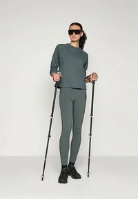 Atletska odjeća u tamnozelenoj boji, koja se sastoji od rebrastog dugog rukava i uskih tajica. Model drži planinarske štapove i nosi crne tenisice.