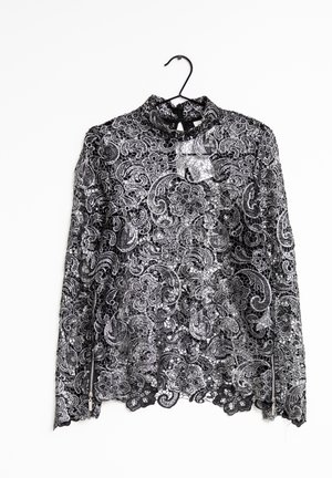 Blouse noire en dentelle florale à manches longues, col montant, détails avec fermeture éclair sur les manches et ourlet festonné, suspendue sur un cintre noir.