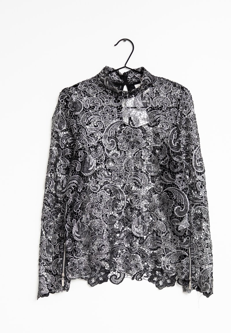 Blouse noire en dentelle florale à manches longues, col montant, détails avec fermeture éclair sur les manches et ourlet festonné, suspendue sur un cintre noir.