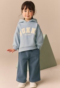 Világoskék kapucnis pulóver, amelyen krémszínű "BUNNY ACADEMY" hímzés található, sötétkék, bő szárú nadrággal és fehér sportcipővel párosítva.