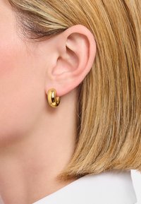 Nahaufnahme eines Ohres einer Frau mit kurzem, blonden Haar, die einen dicken, glänzenden goldenen Creolenohrring vor einem weißen Hintergrund trägt.