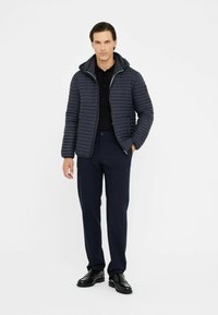 Giubbotto imbottito blu navy con cuciture orizzontali, cappuccio attaccato, zip frontale, abbinato a pantaloni scuri e scarpe nere, su sfondo bianco.
