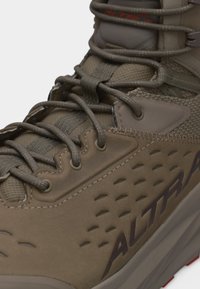 Κοντινό πλάνο ενός καφέ ορειβατικού μποτάκι με λεπτομερείς ραφές, δίχτυ από ύφασμα, στρογγυλά κορδόνια και λογότυπο "ALTRA" στην πλευρά.