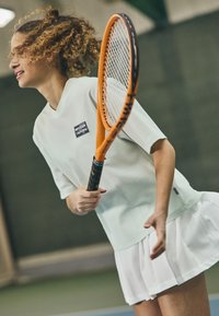 Ανοιχτό πράσινο διχτυωτό μπλουζάκι τένις με χαλαρή εφαρμογή και κοντά μανίκια, συνδυασμένο με μια λευκή πτυχωτή φούστα. Ο παίκτης κρατάει μια πορτοκαλί ρακέτα τένις.