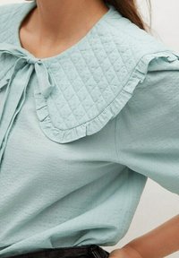 Blouse bleu clair avec un col boutonné matelassé et volanté, et un lien à l'encolure. Le tissu présente un motif texturé subtil.