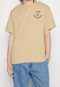 T-shirt en coton beige avec un col rond, des manches courtes et un design graphique arborant "New Balance" et un symbole de main sur le côté gauche de la poitrine.