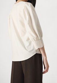 Blusa crema con maniche a palloncino; presenta dettagli in pizzo sui polsini e un collo arrotondato. Indossata con pantaloni larghi marroni scuri.