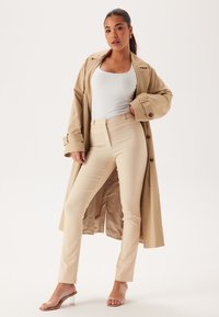 Cappotto trench beige sopra una canotta bianca e pantaloni beige aderenti. Tacchi trasparenti completano l'outfit. Il tessuto appare liscio e ben rifinito.