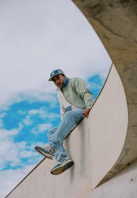 Homme portant une casquette, une veste, un jean et des baskets, assis sur un rebord en béton incurvé devant un ciel bleu avec des nuages.