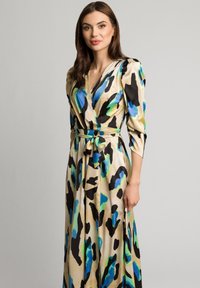 Długa sukienka w stylu wrap w beżowym satynowym materiale, ozdobiona wyrazistymi niebieskimi i zielonymi abstrakcyjnymi wzorami, z rękawami do łokcia i dopasowanym paskiem w talii.