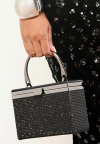 Ruka držící černou obdélníkovou kabelku zdobenou malými lesklými hroty, s kovovým stříbrným držadlem a zapínáním, na pozadí černých flitrových šatů.