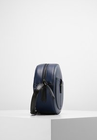 Kulatá crossbody taška z námořnickomodré kůže s černým zipem a nastavitelným popruhem, vystavená na bílém podstavci proti jednoduchému pozadí.