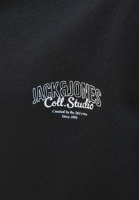 Crna majica s kratkim rukavima s natpisom "JACK & JONES Coll. Studio" u bijelom koncu, s dodatnim redom koji kaže "Kreirali JeJ tim od 1990."
