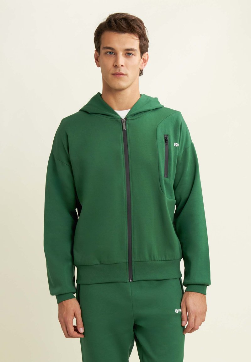 Chaqueta con cremallera verde en tejido suave, que presenta un bolsillo frontal con cremallera, capucha y puños de canalé. Combinada con joggers a juego.