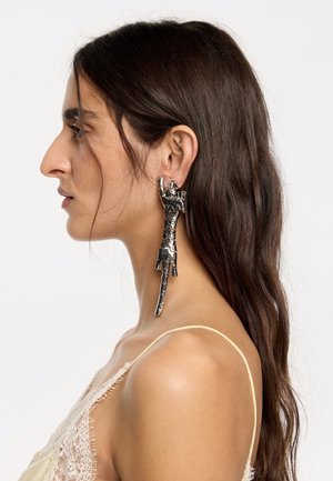 Pendientes grandes en tono plateado con un diseño único que presenta figuras humanas abstractas. Superficie texturizada con acentos brillantes y forma alargada.