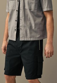 Chemise grise à manches courtes avec deux poches poitrine, associée à un short cargo noir doté d'une poche zippée et de poches latérales à rabat.
