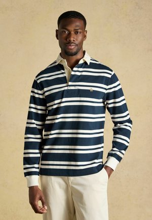 Man wearing navy and white striped long-sleeve rugby shirt with beige collar and light beige pants standing against a plain background.