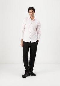 Helles rosa Langarmhemd mit Knopfleiste, kombiniert mit schwarzen Hosen und schwarzen Schuhen, vor einem schlichten weißen Hintergrund.