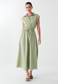 Svetlo zelena obleka do kolen z kratkimi rokavi, spredaj na gumbu, z vrvico za pas in tekočo krilom. Tkanina se zdi mehka in gladka.