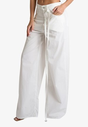 Donna che indossa pantaloni bianchi a vita alta e gamba larga, con bottone frontale, cordino lungo e una mano in tasca, a piedi nudi su sfondo bianco.