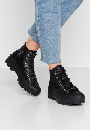Osoba nosząca czarne skórzane wysokie buty Converse z grubymi podeszwami i wzorzystymi sznurowadłami, w połączeniu z jasnoniebieskimi skróconymi dżinsami.