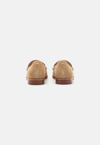 Tan suède instappers met een ronde neus en een lage houten hak. Gladde textuur met minimale stikseldetails. Gouden logo-accenten op de achterkant.