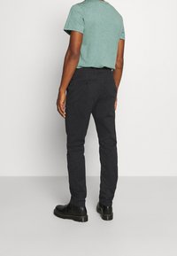 Svarta slim-fit byxor med en slät yta och detaljerad bakficka, tillsammans med en grön t-shirt med kort ärm och svarta ankelstövlar.