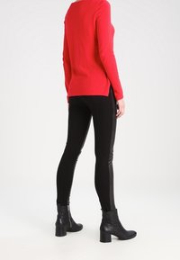 Un pull rouge à manches longues avec des fentes sur les côtés, associé à des leggings noirs avec un panneau latéral brillant et des bottines noires à talon épais.