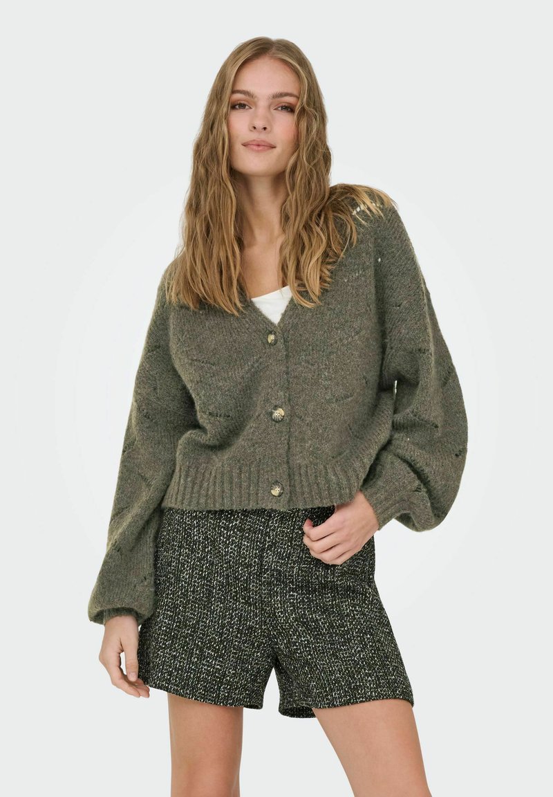 Cardigan verde oliva, oversize, con scollatura a V profonda, polsini e orlo a coste, con tre grandi bottoni. Abbinato a pantaloni corti neri strutturati.