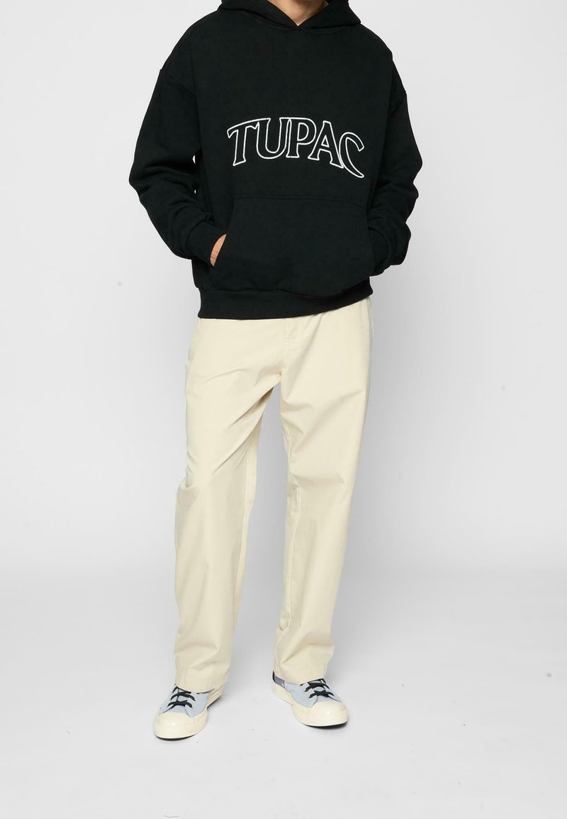 Svart huvtröja med vit text "TUPAC", kombinerad med krämfärgade byxor och grå sneakers med svarta snören och vita sulor.