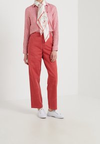 Osoba oblečená v červeném pruhovaném tričku, červených kalhotách s vysokým pasem, bílém květinovém šálu a bílých teniskách stojící na jednobarevném pozadí.